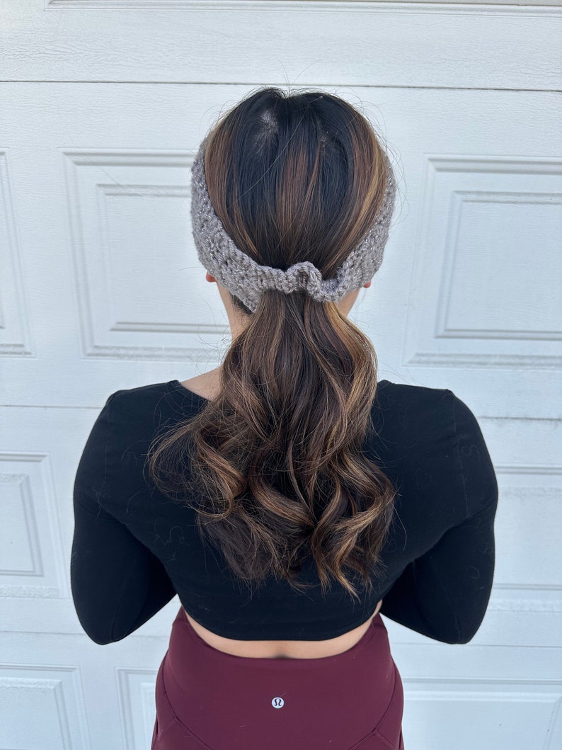 May include: A gray knitted headband worn by a person with long brown hair pulled back in a ponytail. The headband is tied in a knot at the back of the head.
