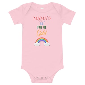 Peut inclure: Body rose clair pour bébé avec l'inscription "Mama's Lil' Pot of Gold" en rouge, vert et or. Un arc-en-ciel coloré avec des nuages blancs est en dessous. Le body a des manches courtes et des boutons-pression en bas.