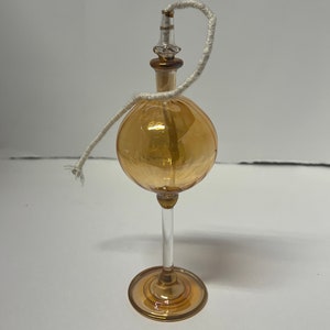 May include: A clear glass oil lamp with a round amber-colored body and a clear glass stem. The lamp has a white wick and a gold-colored metal top.