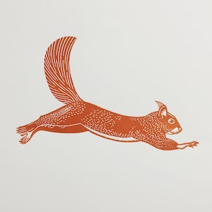 May include: A linocut print of a squirrel in mid-leap, rendered in shades of orange. The squirrel's body is detailed with small dots and lines, and its bushy tail curves upwards. The background is plain white.