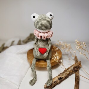 May include: A crocheted green frog toy holding a red heart. The frog is wearing a pink ruffled collar and has white eyes. The frog is sitting on a wooden base.