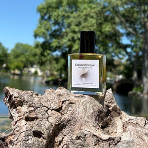 May include: A rectangular glass bottle with a black cap, labelled "Karmic Reversal return to sender oil". The bottle sits on a textured piece of wood, with a lake and trees in the background. The label features a yin-yang symbol.