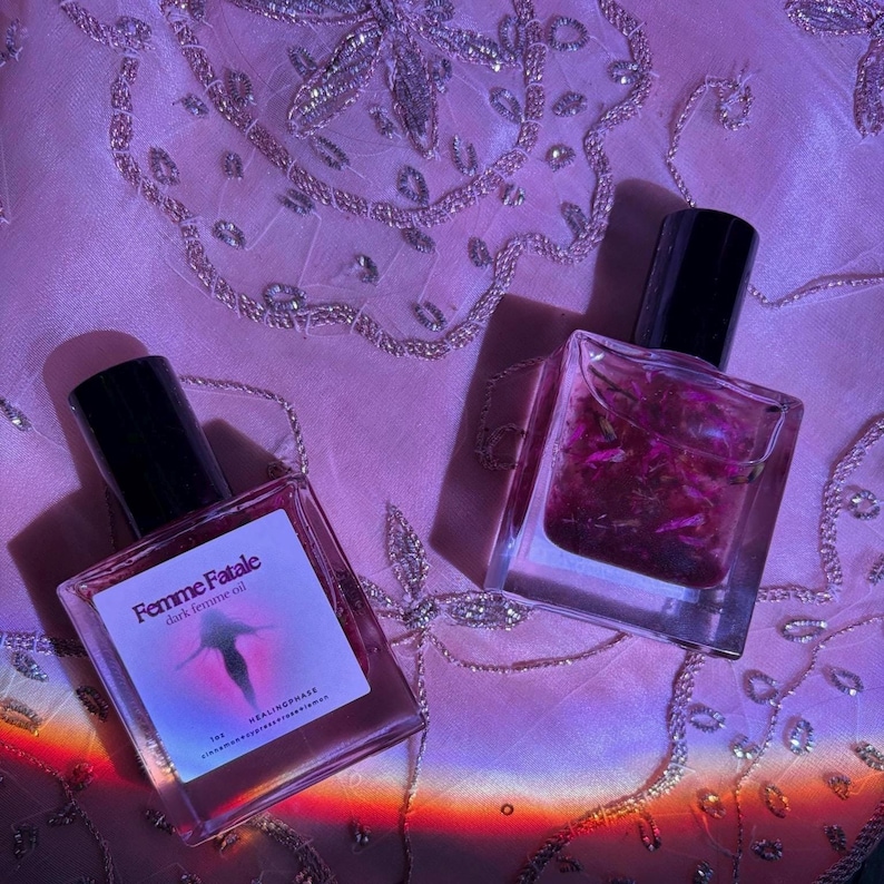 May include: Two glass bottles of dark femme oil with black caps sit on a pink, embroidered surface. One bottle has a label with the text "Femme Fatale" and a silhouette. The other bottle contains pink petals.
