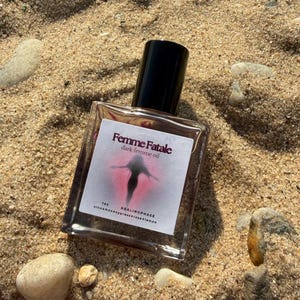 May include: A clear glass bottle of "Femme Fatale dark femme oil" with a black cap, resting on sand. The label features a pink and black graphic. The bottle is surrounded by small rocks and sand.