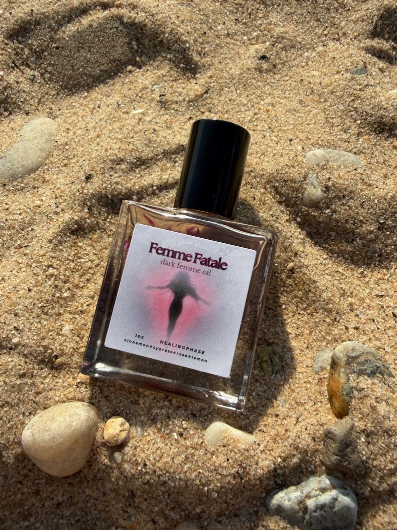 May include: A clear glass bottle of "Femme Fatale dark femme oil" with a black cap, resting on sand. The label features a pink and black graphic. The bottle is surrounded by small rocks and sand.