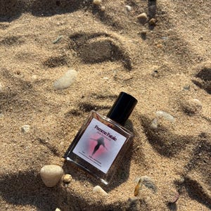 May include: A clear glass perfume bottle with a black cap, resting on sand. The label reads "Femme Fatale" with a graphic of a figure. The bottle is surrounded by small rocks and pebbles, with the sand in the background.