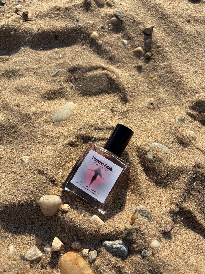 May include: A clear glass perfume bottle with a black cap, resting on sand. The label reads "Femme Fatale" with a graphic of a figure. The bottle is surrounded by small rocks and pebbles, with the sand in the background.