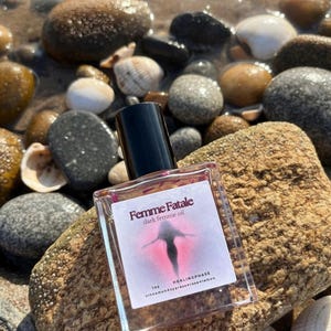 May include: A clear glass bottle of "Femme Fatale dark femme oil" with a black cap, resting on a textured rock. The label features a silhouette and the words "HEALINGPHASE". The background includes pebbles, shells, and water.