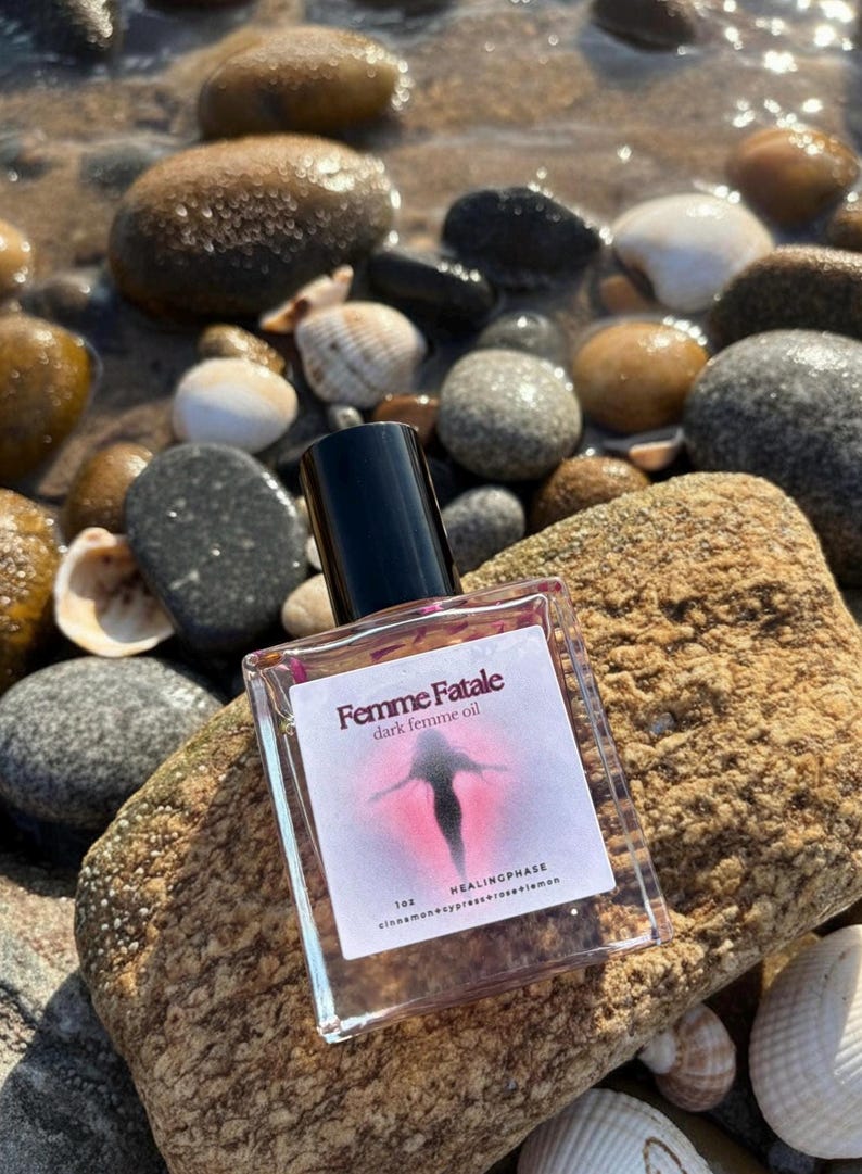 May include: A clear glass bottle of "Femme Fatale dark femme oil" with a black cap, resting on a textured rock. The label features a silhouette and the words "HEALINGPHASE". The background includes pebbles, shells, and water.