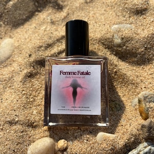 May include: A clear glass bottle with a black cap, labeled "Femme Fatale dark femme oil." The label features a silhouette of a person in a pink and red design. The bottle is on a sandy surface with small rocks.
