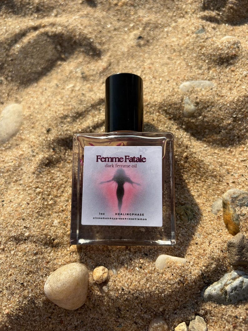 May include: A clear glass bottle with a black cap, labeled "Femme Fatale dark femme oil." The label features a silhouette of a person in a pink and red design. The bottle is on a sandy surface with small rocks.