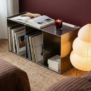 Può includere: Un moderno mobile contenitore in metallo con scomparti aperti, contenente libri e dischi in vinile. Un libro aperto e una candela accesa sono in cima. Una lampada a lanterna di carta è sullo sfondo.
