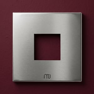 Op de afbeelding: Een vierkante, geborstelde metalen wandplaat met een centraal vierkant gat. De plaat is bevestigd aan een dieprode muur. In de rechter benedenhoek staat een klein, gestileerd logo.