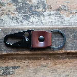 May include: A black metal key chain with a brown leather strap and a snap closure. The key chain has a round metal ring.