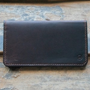 May include: A dark brown leather wallet with a stitched border and a small embossed logo in the lower right corner. The wallet is rectangular and appears to be closed. The background is a weathered wooden surface.