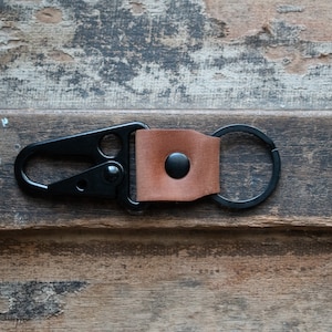May include: A black metal key chain with a brown leather loop and a metal ring. The key chain is on a wooden surface.