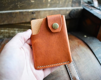 May include: A small, rectangular, brown leather card holder with a flap secured by a brass snap. The card holder is held in a hand, showcasing its compact design and stitched edges. The leather has a natural, textured appearance.