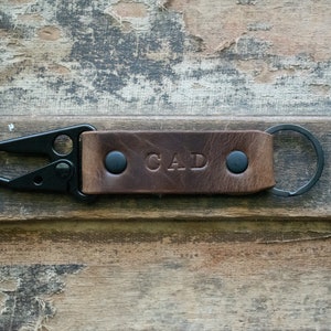 Könnte beinhalten: Brauner Leder-Schlüsselanhänger mit einem schwarzen Metall-Karabinerhaken und einem Schlüsselring. Das Leder ist mit den Initialen "CAD" geprägt.