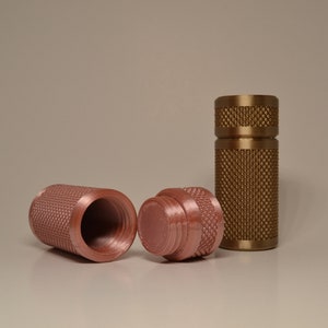 Op de afbeelding: Drie metalen containers met een getextureerd oppervlak. Twee zijn licht koperkleurig en één is goudkleurig. De koperen containers zijn open en de gouden container is gesloten.