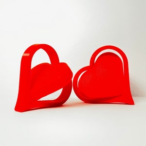 May include: Two red heart-shaped cookie cutters. The cookie cutters are made of plastic and are ready to be used to make heart-shaped cookies.