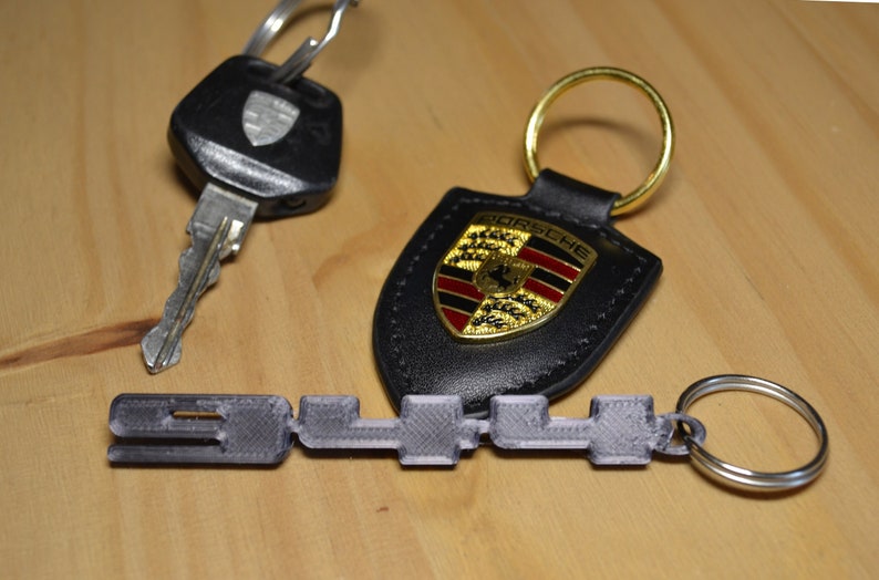 May include: A set of three keyrings. One keyring is a black leather shield with a gold Porsche logo. Another keyring is a silver metal ring with a 3D printed black plastic key shaped like the word "911". The third keyring is a black key with a silver metal blade and a black plastic head with a silver Porsche logo.