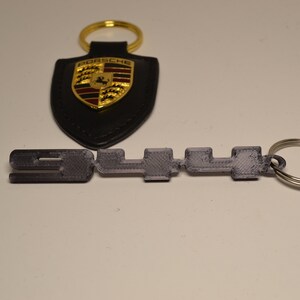 May include: A black leather Porsche keyring with a gold shield logo and the text "PORSCHE". A silver metal keyring with the text "911" is attached to the leather keyring.