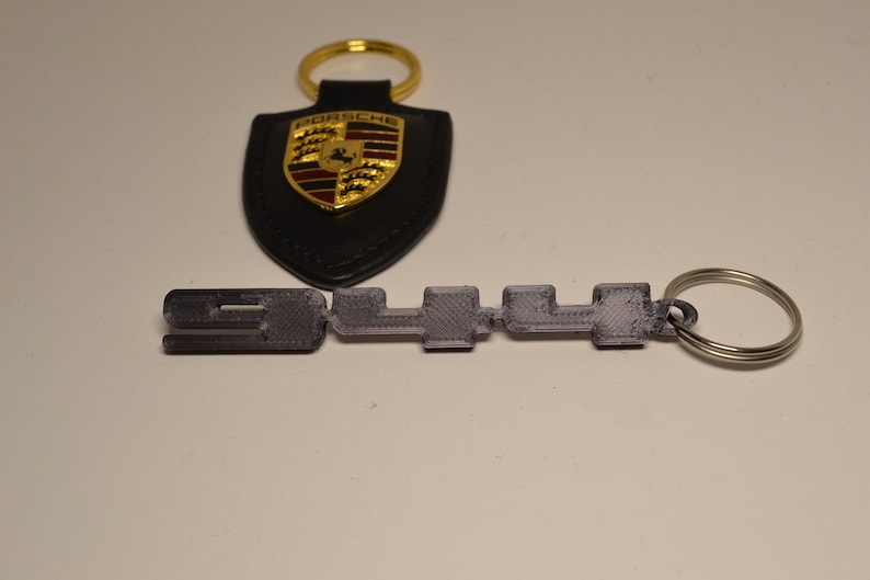 May include: A black leather Porsche keyring with a gold shield logo and the text "PORSCHE". A silver metal keyring with the text "911" is attached to the leather keyring.