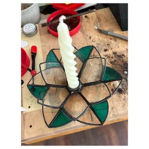 May include: A handcrafted stained glass candle holder in a flower shape. The holder features clear and emerald green glass panels, framed by black solder. A white, spiral-shaped candle sits in the center, ready to be lit.