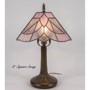 May include: Cleopatra Pendant Lamp, 8" Square Swag. A table lamp with a bronze-toned metal base and a textured, light pink stained glass shade featuring a geometric petal pattern. The lamp emits a warm glow from the center of the shade.