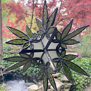 May include: A stained glass suncatcher with a design of green leaves. The suncatcher is hanging from a string and is in front of a blurry background of trees.