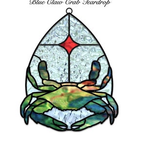 May include: A stained glass teardrop-shaped ornament featuring a blue crab with green and brown markings. The ornament has a white background with a red diamond in the center.