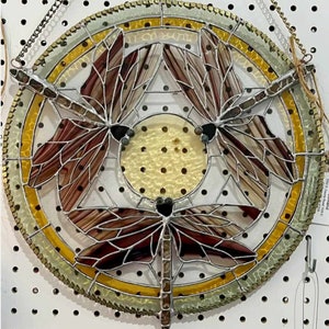 May include: A stained glass suncatcher featuring three dragonflies with brown and white wings. The dragonflies are arranged in a circle with a yellow and green border.