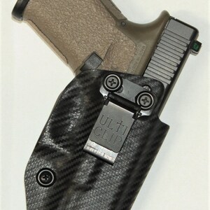 Puede incluir: Una funda negra y gris con un patr&oacute;n de fibra de carbono y un Ulti-Clip negro. Una pistola de color beige est&aacute; guardada en la funda.