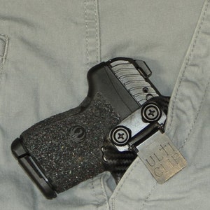 Puede incluir: Una pistola negra con un agarre texturizado y un Ulti-Clip plateado fijado en el lateral. La pistola est&aacute; metida en el bolsillo de un pantal&oacute;n gris.