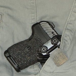 May include: A black handgun with a textured grip and a silver Ulti-Clip attached to the side. The gun is tucked into the pocket of a pair of grey trousers.