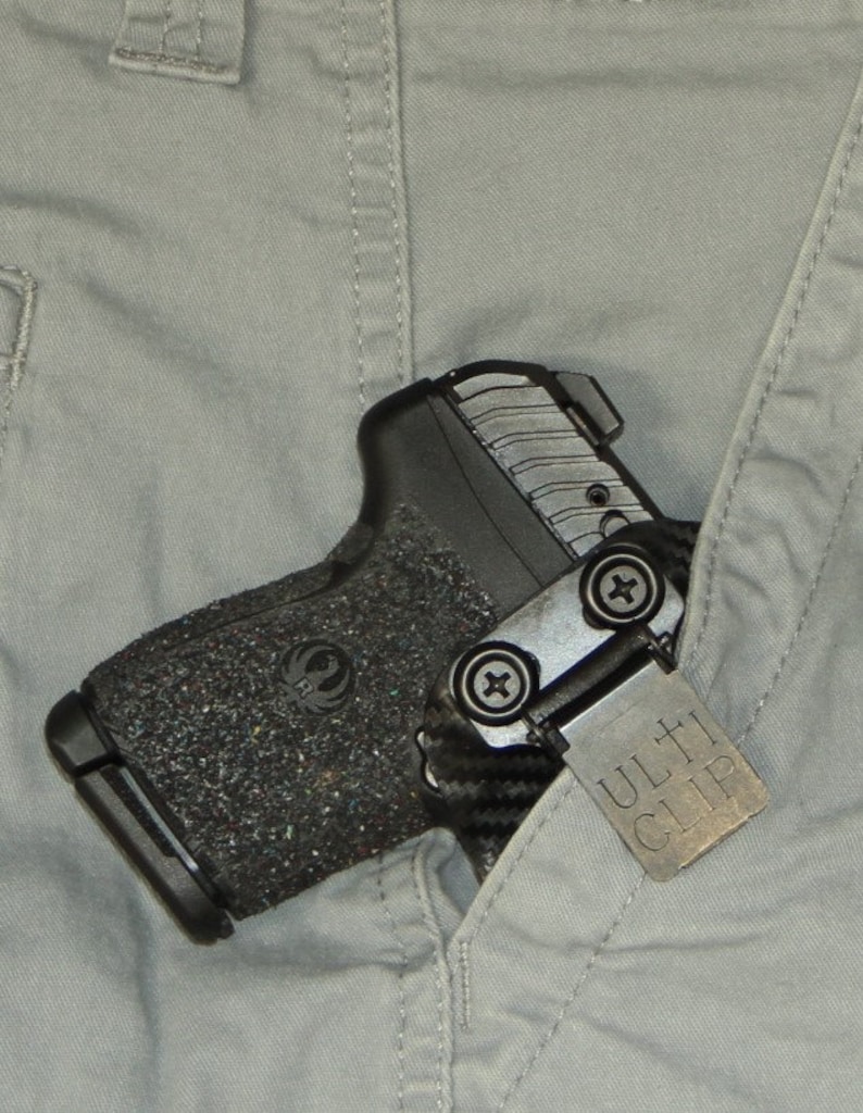 May include: A black handgun with a textured grip and a silver Ulti-Clip attached to the side. The gun is tucked into the pocket of a pair of grey trousers.