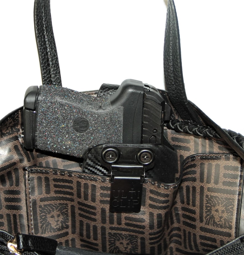 May include: A black handgun with a black holster is secured inside a brown handbag with a black handle. The holster has a black clip with the text "ULTI-CLIP" printed on it.