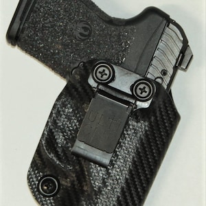 May include: A black and grey gun holster with a carbon fibre pattern and a black Ulti-Clip. The holster has a screw on the side and a metal clip.