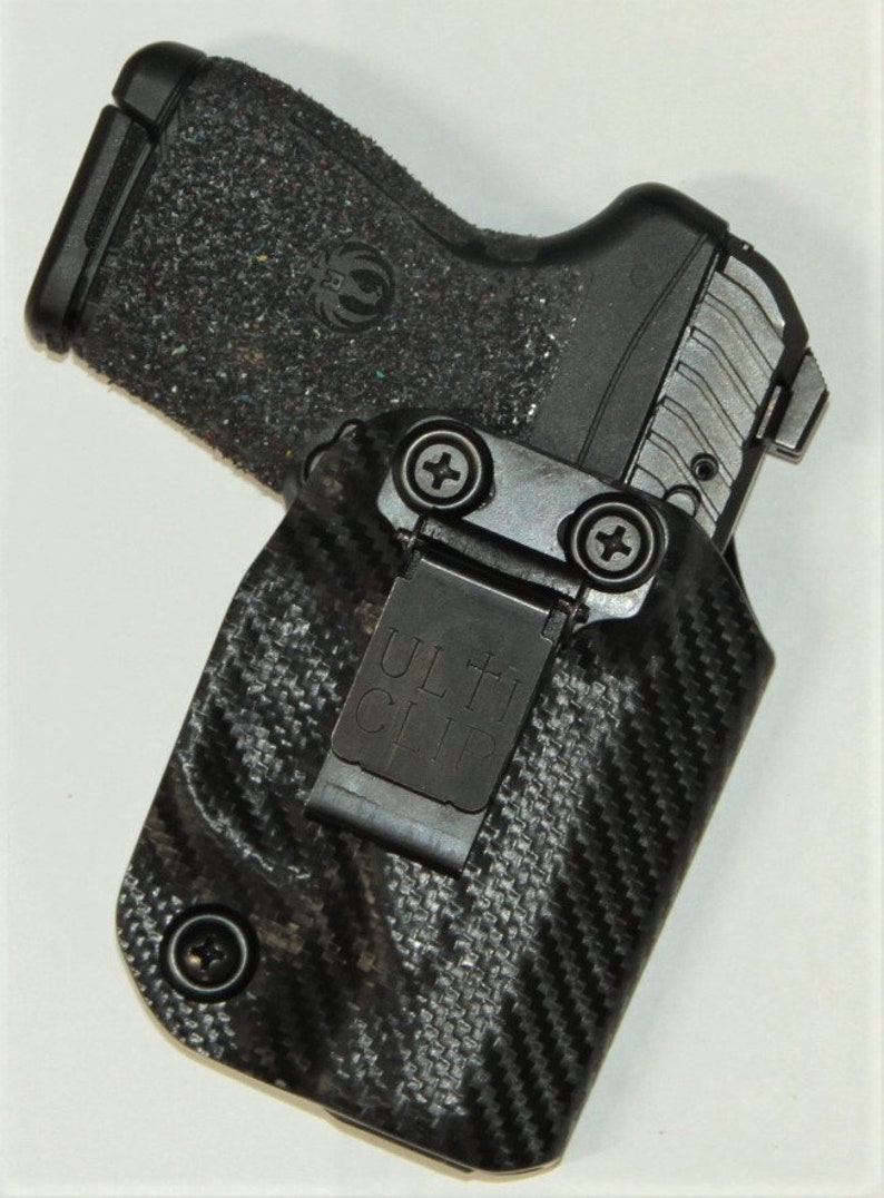 May include: A black and grey gun holster with a carbon fibre pattern and a black Ulti-Clip. The holster has a screw on the side and a metal clip.