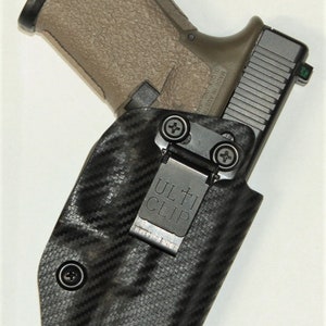 May include: A black and grey holster with a carbon fibre pattern and a black Ulti-Clip. A tan handgun is holstered in the holster.