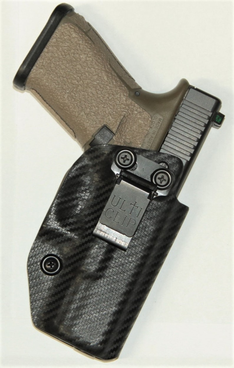 May include: A black and grey holster with a carbon fibre pattern and a black Ulti-Clip. A tan handgun is holstered in the holster.