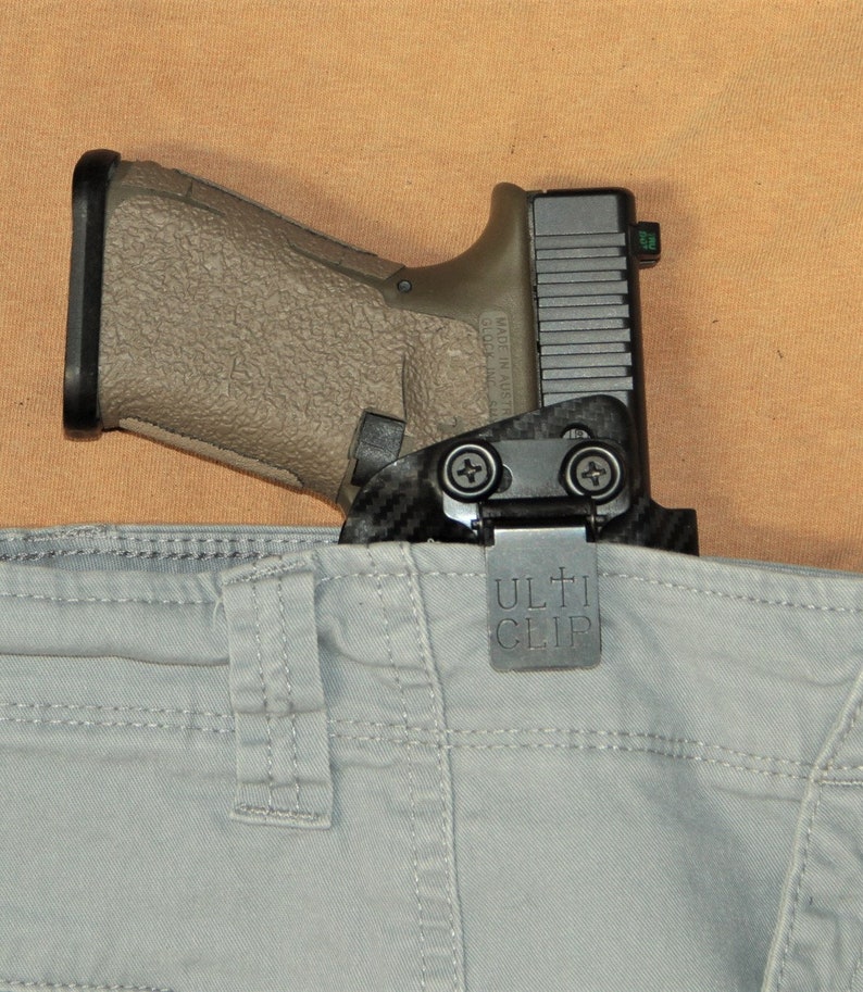 Puede incluir: Una funda para pistola negra y gris con un patr&oacute;n de fibra de carbono y un clip de metal que dice "ULTI CLIP". La funda est&aacute; unida a un pantal&oacute;n gris.