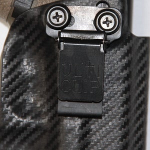 May include: A black holster with a carbon fibre pattern and a black metal clip. The clip has the word "ULTI CLIP" printed on it.