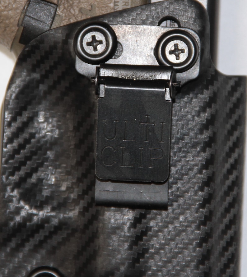 May include: A black holster with a carbon fibre pattern and a black metal clip. The clip has the word "ULTI CLIP" printed on it.