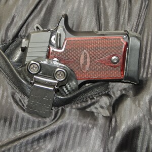 May include: A black handgun with a red wooden grip is tucked into a black fabric holster with a black clip that says "ULTI-CLIP".