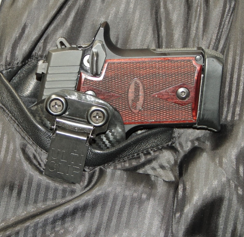 May include: A black handgun with a red wooden grip is tucked into a black fabric holster with a black clip that says "ULTI-CLIP".