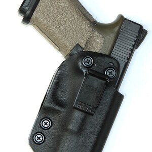 Puede incluir: Una funda de cuero negra con un clip y dos tornillos. Una pistola negra y marr&oacute;n est&aacute; dentro de la funda.