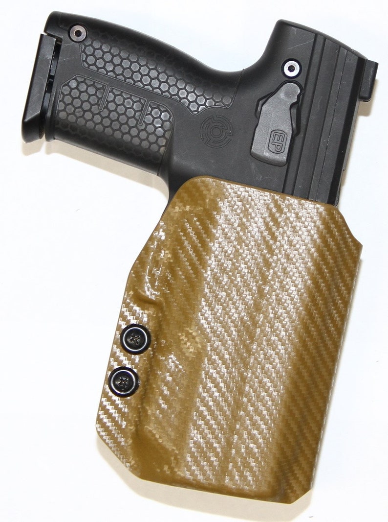 Puede incluir: Una funda de cuero marr&oacute;n con un patr&oacute;n de fibra de carbono y broches negros. La funda est&aacute; dise&ntilde;ada para sostener una pistola. La funda tiene un guardamonte de pl&aacute;stico negro.