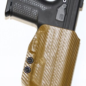 May include: A tan leather holster with a carbon fibre pattern and black snaps. The holster is designed to hold a handgun. The holster has a black plastic trigger guard.