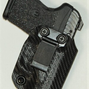 Puede incluir: Una funda para pistola negra y gris con un patr&oacute;n de fibra de carbono y un Ulti-Clip negro. La funda tiene un tornillo en el lateral y un clip de metal.